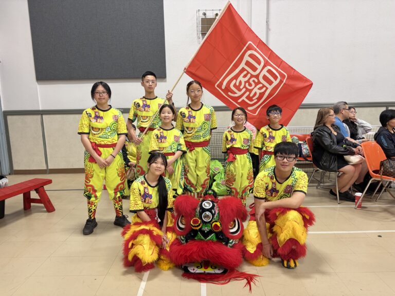 8 Chinese lion dance team members holding a big red flag with the KSK logo posing with a red and green Chinese lion.