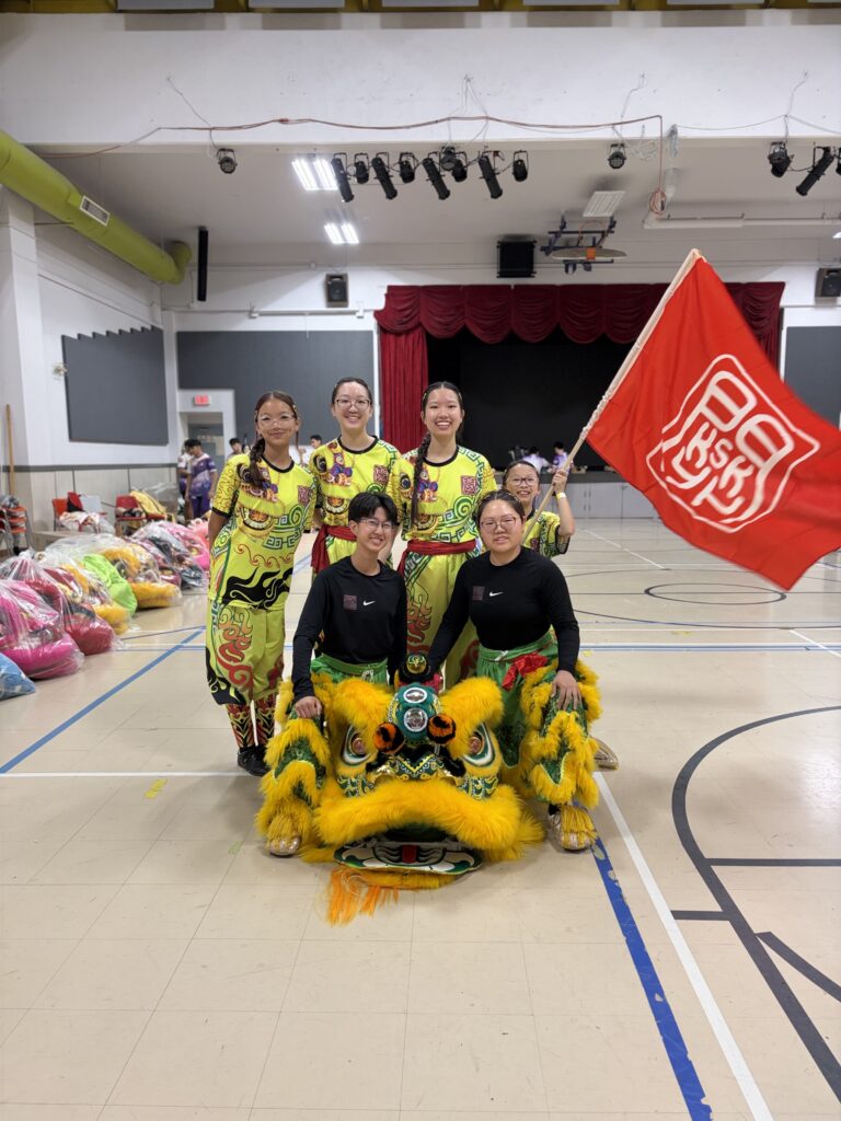 6 all female Chinese lion dance team members holding a big red flag with the KSK logo posing with a yellow and green Chinese lion.