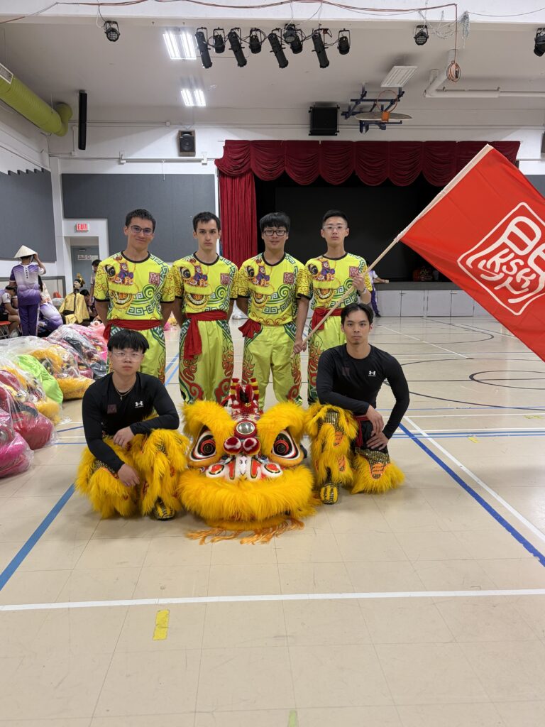 6 all male Chinese lion dance team members holding a big red flag with the KSK logo posing with an orange and yellow colored Chinese lion.